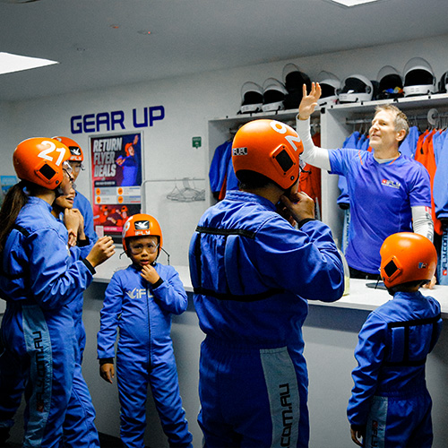 An instructor briefing a group wearing blue flight suits and orange helmets